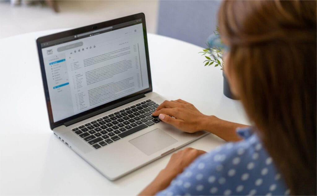 The image features a woman in a light blue, polka dot shirt using a laptop. The laptop screen displays an email client or content management system with various options and tabs visible, suggesting professional or personal productivity tasks such as managing communications or scheduling. The setting appears to be a clean and modern workspace, perhaps a home office or a co-working space, indicated by the casual clothing and the presence of a potted plant, which adds a touch of warmth and personalization to the environment. The focus on the laptop screen and the woman’s concentrated expression highlight her engagement and attentiveness to the task at hand. This setting is typical for professionals who manage their tasks digitally, reflecting a common contemporary workflow in many fields.