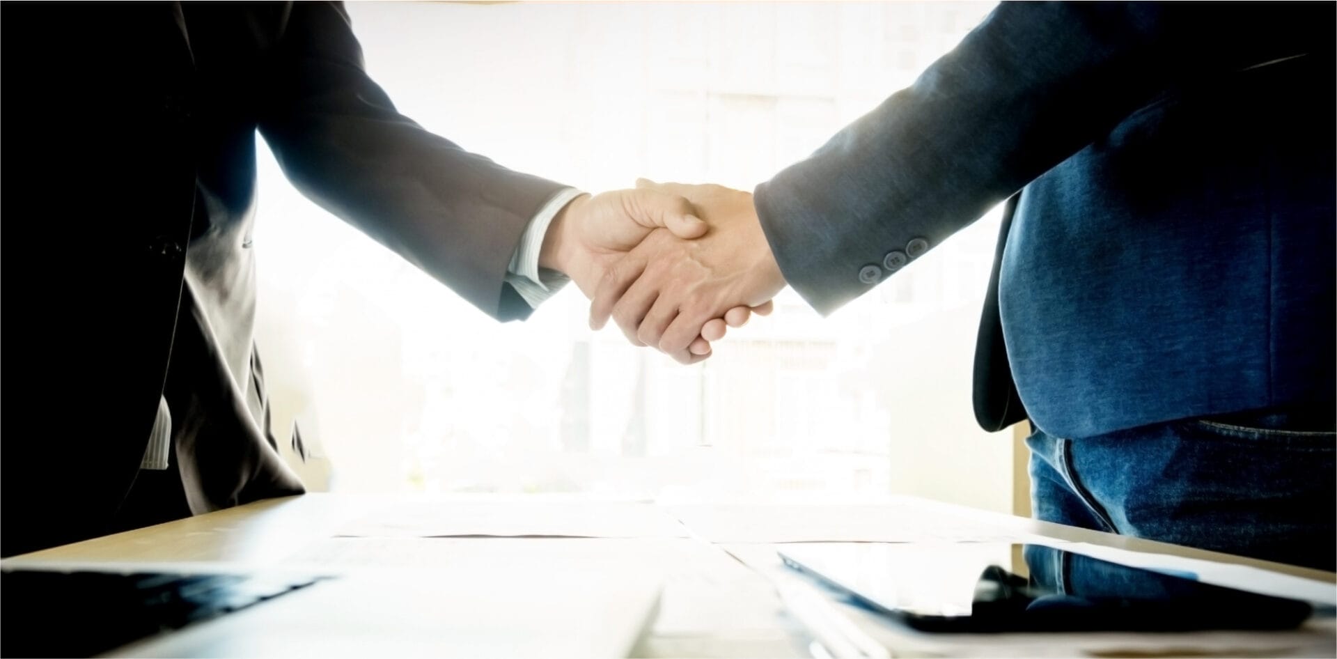 The image features two business professionals shaking hands across a table, symbolizing a successful agreement or partnership. This kind of imagery is commonly used to represent business deals, collaborations, or the closure of negotiations. The backlighting and focus on the handshake emphasize the positive outcome and mutual agreement in a professional setting.