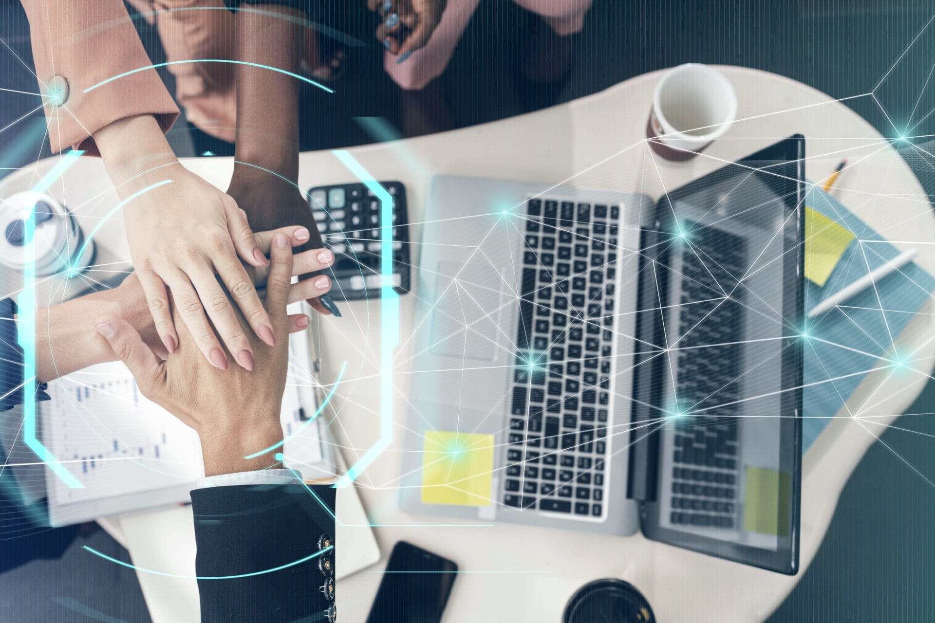 This image shows a team of professionals stacking hands over a worktable, symbolizing unity and teamwork, with a digital overlay of network connections, emphasizing collaboration in a modern, tech-driven environment.