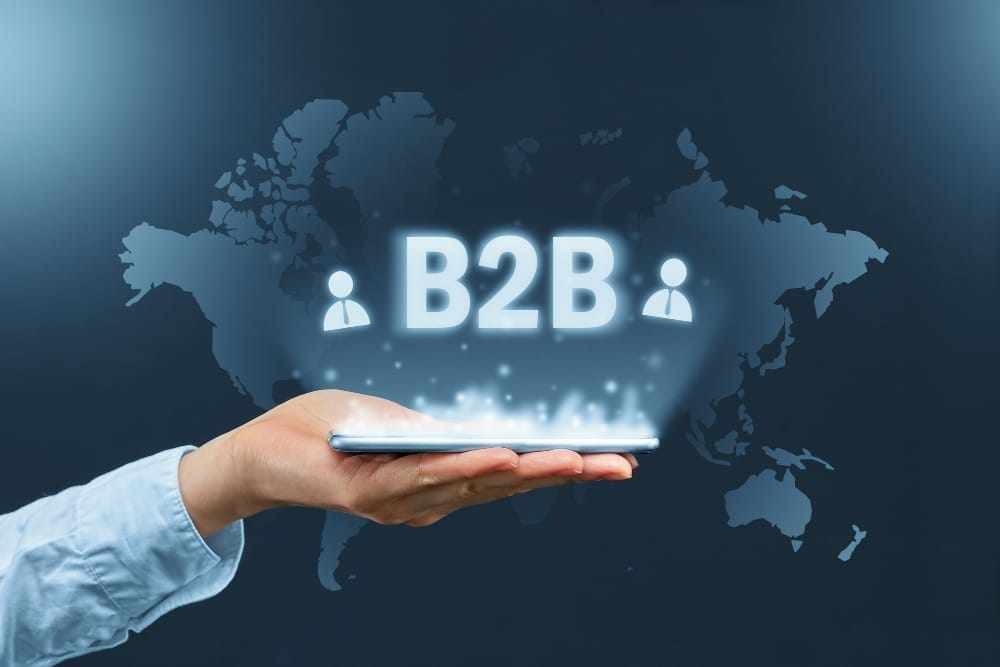 A hand holding a smartphone with the glowing text "B2B" and icons of two people above it. The background features a world map in a dark blue hue, highlighting the global aspect of B2B interactions.